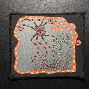 May include: A rectangular black patch with a gray background featuring a stylized creature design. The creature has a red and black body with long, thin legs. The patch is outlined in orange and yellow.