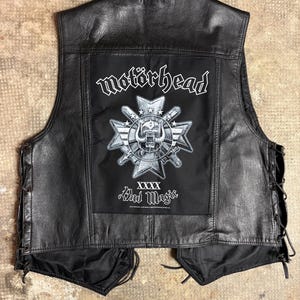 Op de afbeelding: Zwarte leren vest met het logo van de band Motörhead op de rug. Het logo bevat de bandnaam, een schedel en de woorden "Bad Magic". Het vest heeft veters aan de zijkanten en een klassieke biker-stijl.