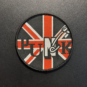 Könnte beinhalten: Ein runder, bestickter Aufnäher mit einem Union-Jack-Flaggenmotiv in Schwarz, Weiß und Rot. Das Wort PUNK ist in der Mitte in fetten schwarzen und weißen Buchstaben gestickt, wobei eine stilisierte Sicherheitsnadel in den Buchstaben N integriert ist.