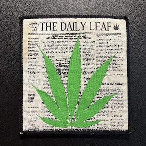 Op de afbeelding: Een rechthoekige stoffen patch met een zwarte gestikte rand. De achtergrond toont een witte krantenprint met de tekst THE DAILY LEAF bovenaan. Een grote, felgroene cannabisblad-afbeelding is gecentreerd over de gedrukte tekst.