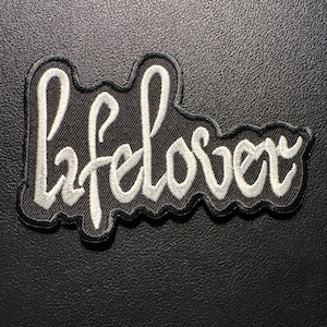 May include: A black patch with the white embroidered word "lifelover". The patch has a black border and is set against a dark background. The lettering is in a cursive style.