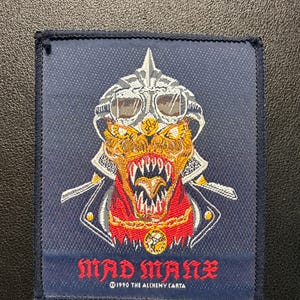 Könnte beinhalten: Ein rechteckiger gestickter Aufnäher auf dunkelblauem Grund mit einer Kreatur, die einen silbernen Helm mit Schutzbrille trägt. Die Kreatur hat spitze Zähne und einen roten Halsbereich. Der Text MAD MANX und 1990 THE ALCHEMY CARTA ist rot gestickt.