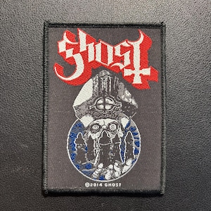Könnte beinhalten: Schwarzer rechteckiger Aufnäher mit dem Wort "GHOST" in roten und weißen gotischen Buchstaben. Darunter eine detaillierte Illustration eines Schädels mit Hut, umgeben von einem blauen Kreis. Der Text "©2014 GHOST" ist unten aufgedruckt.