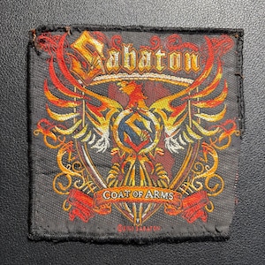 Könnte beinhalten: Gestickter Aufnäher mit dem Wort "Sabaton" über einem stilisierten Adler. Der Aufnäher hat einen schwarzen Hintergrund mit roten, orangefarbenen und gelben Details. Der Text "COAT OF ARMS" ist ebenfalls sichtbar.