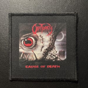 Peut inclure: Un écusson carré en tissu noir présentant une impression graphique de la pochette de l'album Cause of Death d'Obituary. Le motif comprend un grand œil détaillé avec un iris rouge, des éléments texturés sombres et le texte Obituary en rouge.