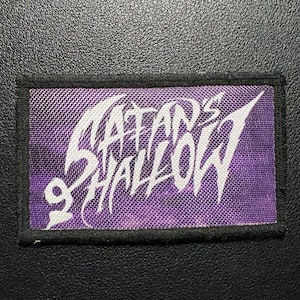 May include: A rectangular patch with the words "SATAN'S SHALLOW" in white, stylized lettering on a purple background. The patch has a black border and a small symbol. The patch is likely for clothing or accessories.