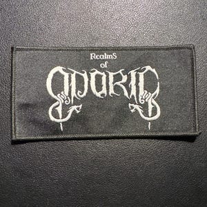 Może przedstawiać: Czarna prostokątna naszywka z białym haftowanym tekstem. Tekst brzmi "Realms of ODORIC" w stylizowanej czcionce. Naszywka jest prawdopodobnie przeznaczona na odzież lub akcesoria.
