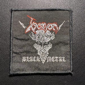 Könnte beinhalten: Schwarzer quadratischer Aufnäher mit dem Wort "Venom" in roter und silberner gotischer Schrift, über einem silbernen Totenkopf-Design. Die Worte "BLACK METAL" stehen unter dem Totenkopf in silberner gotischer Schrift.