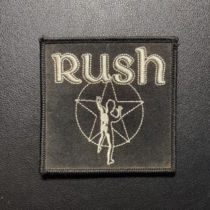 以下が含まれることがあります： 上部に白い糸で「RUSH」という文字が刺繍された、正方形の黒い布製ワッペン。テキストの下には、白い刺繍で星のシンボルが描かれており、その中に腕を上げた様式化された人物像が白いステッチで縁取られています。