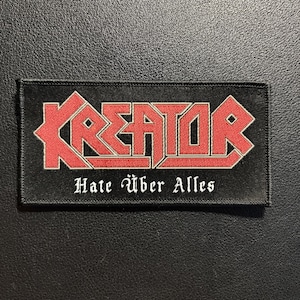 Könnte beinhalten: Ein rechteckiger schwarzer Stoffaufnäher mit dem rot-weiß gestickten Schriftzug KREATOR in einer stilisierten Schriftart. Darunter sind die Worte Hate Über Alles in weißer, gotisch anmutender Schrift auf dem schwarzen Hintergrund gestickt.