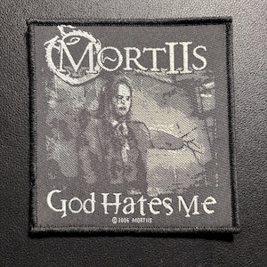 Könnte beinhalten: Schwarz-weißer Mortiis-Aufnäher mit dem Text "MORTIIS" oben und "God Hates Me" unten. Der Aufnäher zeigt ein Graustufenbild einer Person. Der Text "©2006 MORTIIS" ist darunter gedruckt.