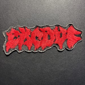 Könnte beinhalten: Ein roter, gestickter Aufnäher mit dem Wort "BROKE" in einer stilisierten Schriftart. Der Aufnäher hat einen schwarzen Rand und eine transparente Rückseite. Er liegt auf einer schwarzen, strukturierten Oberfläche.