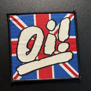 May include: A square embroidered patch featuring the Union Jack flag in red, white, and blue. The word Oi! is stitched in large, bold white letters across the center of the flag. The entire patch is bordered by a thick black stitched edge.
