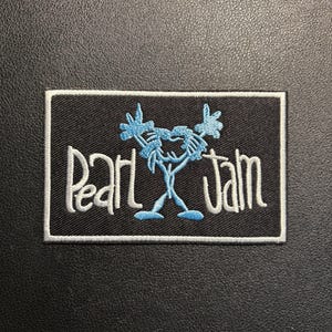 Puede incluir: Parche rectangular negro con borde blanco. El parche presenta el nombre de la banda "Pearl Jam" en escritura blanca, con un diseño azul de figura de palo sobre el texto. El parche es probablemente para ropa o accesorios.