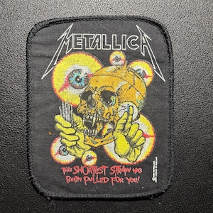 Könnte beinhalten: Schwarzer, rechteckiger Aufnäher mit einem gelben Totenkopf und weißem Metallica-Logo. Der Totenkopf ist von gelben und roten Augäpfeln umgeben. Der Text "THIS SHORTEST STRAW HAS BEEN PULLED FOR YOU!" ist rot gedruckt.