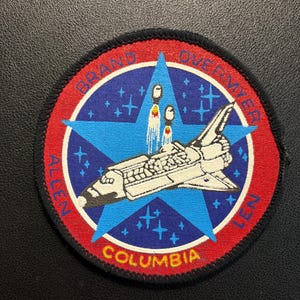 Könnte beinhalten: Ein runder bestickter Aufnäher mit einer Weltraumfähre im Flug vor einem blauen Stern und einem roten Rand. Der Text lautet BRAND, OVERMYER, ALLEN, LEN und COLUMBIA. Das Design enthält kleine weiße Sterne auf dunkelblauem Hintergrund.