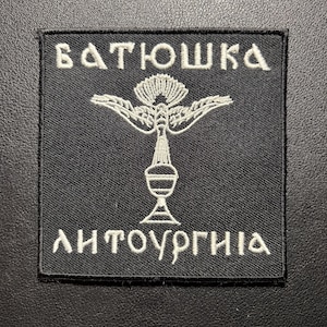 May include: A square black embroidered patch featuring white thread detailing. The design includes a central chalice topped with a bird, flanked by the Cyrillic text "БАТЮШКА" above and "ЛИТУРГІЯ" below, all enclosed within a stitched black border.