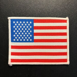 Könnte beinhalten: Ein rechteckiger bestickter Aufnäher mit der Flagge der Vereinigten Staaten von Amerika. Das Design zeigt ein blaues Feld mit weißen Sternen und abwechselnd horizontalen roten und weißen Streifen, alles umrandet von einer weißen Kante.