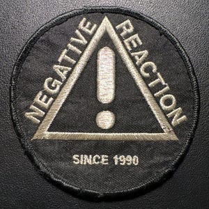 Könnte beinhalten: Schwarzer runder Aufnäher mit den Worten "NEGATIVE REACTION" um ein weißes Dreieck mit einem Ausrufezeichen. Der Text "SINCE 1990" steht darunter. Gestickt mit weißem Faden.