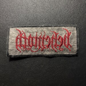 K&ouml;nnte beinhalten: Ein rechteckiger Stoffaufn&auml;her mit einer grauen, filzartigen Basis und einem schwarzen, gestickten Rand. In der Mitte befindet sich ein komplexer, stilisierter Schriftzug in leuchtend rotem Garn, der durch spitze, l&auml;ngliche und geschwungene Buchstaben besticht.