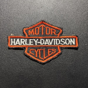 Può includere: Una toppa ricamata Harley-Davidson Motor Cycles a forma di scudo nero con bordo arancione brillante. Il testo MOTOR si trova in alto, CYCLES in basso e HARLEY-DAVIDSON è scritto in lettere bianche su una barra orizzontale arancione.