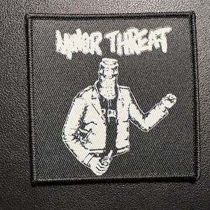 Peut inclure: Écusson brodé noir et blanc avec les mots "MINOR THREAT" dans une police stylisée. L'écusson présente un graphisme d'une personne en veste tenant une bouteille. L'écusson est carré avec une bordure noire.