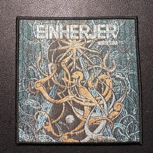 Könnte beinhalten: Quadratischer Aufnäher mit dem Bandnamen "EINHERJER" und dem Text "NORTH STAR". Der Aufnäher zeigt eine detaillierte Illustration in Gold-, Türkis- und Silbertönen mit einem schwarzen Rand. Das Design umfasst komplizierte Muster und ein zentrales sternförmiges Element.