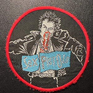 May include: Round black patch with a red border featuring a punk rock illustration. The image depicts a figure with spiky hair and a bloody mouth, holding a sign that reads "Sex Pistols" in blue letters.