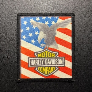Könnte beinhalten: Ein rechteckiger Stoffaufnäher mit einer Grafik eines Weißkopfseeadlers im Flug vor dem Hintergrund der amerikanischen Flagge. Unter dem Adler befindet sich ein schildförmiges Emblem mit dem Text MOTOR HARLEY-DAVIDSON COMPANY in Gelb und Weiß.