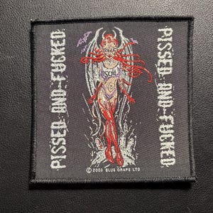 Pode incluir: Patch preto bordado com as palavras "PISSED AND FUCKED" na parte superior e inferior. O centro apresenta uma mulher diabo ruiva com asas. O patch é de 2003.