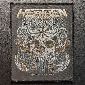 May include: A rectangular embroidered patch featuring a dark gray fabric base with the word HEATHEN stitched in metallic thread at the top. The design includes a central skull with horns, a star symbol, and intricate patterns in silver and bronze.