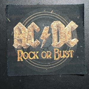 Könnte beinhalten: Schwarzer rechteckiger Aufnäher mit dem Bandnamen "AC/DC" in einem strukturierten, goldfarbenen Schriftzug und einem Blitz-Grafik. Darunter stehen die Worte "ROCK OR BUST" in einer ähnlichen goldenen Schrift. Das Design hat einen schwarzen Hintergrund.