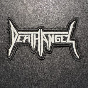 Könnte beinhalten: Gestickter Aufnäher mit dem Bandnamen "Death Angel" in weißer, stilisierter Schrift und schwarzem Rand. Der Aufnäher hat eine rechteckige Form mit spitzen Kanten, vor einem strukturierten schwarzen Hintergrund.