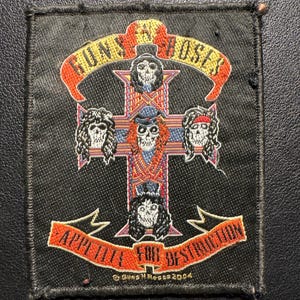 Könnte beinhalten: Ein schwarzer quadratischer Aufnäher mit dem Guns N' Roses-Logo. Das Design zeigt ein Kreuz mit Totenkopfgesichtern, den Bandnamen in Orange und Gelb und den Schriftzug "Appetite for Destruction" in Orange. Der Aufnäher hat einen schwarzen Rand.