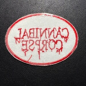 May include: An oval-shaped patch with a white background and a red border. The text "CANNIBAL CORPSE" is embroidered in red, with the letters appearing in a dripping, stylized font. The patch is likely for clothing or accessories.