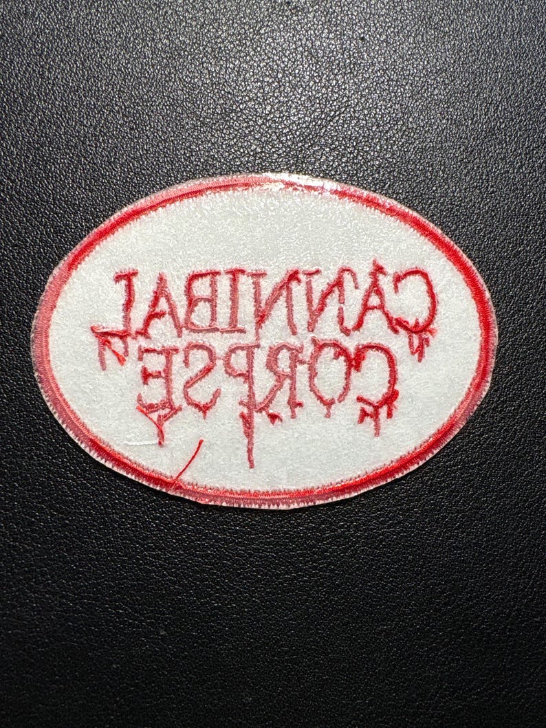 May include: An oval-shaped patch with a white background and a red border. The text "CANNIBAL CORPSE" is embroidered in red, with the letters appearing in a dripping, stylized font. The patch is likely for clothing or accessories.