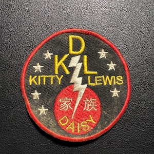 Könnte beinhalten: Runder, gestickter Aufnäher mit schwarzem Hintergrund, rotem Rand und gelber Schrift. Der Aufnäher zeigt den Text "D K L KITTY LEWIS", einen Blitz und einen roten Kreis mit chinesischen Schriftzeichen und dem Wort "DAISY".