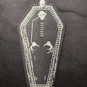Könnte beinhalten: Ein schwarz-weißer, gestickter Aufnäher mit einem Vampir in einem Sarg. Der Vampir hat spitze Zähne und lange Finger. Der Sarg ist weiß umrandet und hat einen dekorativen Rand. Oben auf dem Sarg befindet sich ein Totenkopf.