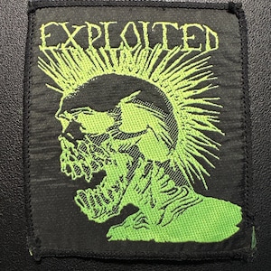 Pode incluir: Patch quadrado preto com a palavra "EXPLOITED" em verde néon acima de um gráfico de caveira verde com moicano. O patch tem uma borda preta e é projetado para ser costurado em roupas.