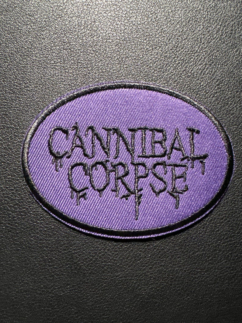 May include: An oval patch featuring the words "CANNIBAL CORPSE" in black, gothic lettering on a purple background. The patch has a black border and is set against a black textured surface.