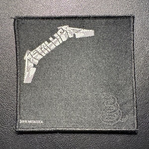 May include: A black square patch with the band name "METALLICA" in silver, stylized lettering. The patch also features a coiled snake emblem and the text "©1991 METALLICA".