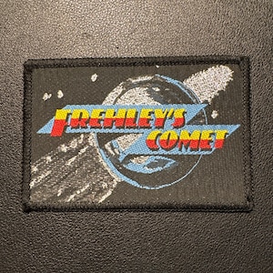 Puede incluir: Un parche rectangular con las palabras "FREHLEY'S COMET" en letras rojas, amarillas y azules. El parche presenta un diseño de cometa sobre un fondo negro. El parche tiene un borde negro.