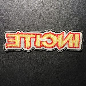 Op de afbeelding: Geborduurde patch met het woord "HUSTLE" in geel, omrand met rood en een witte rand. De patch heeft een rechthoekige vorm en is geplaatst tegen een zwarte achtergrond. De tekst is in een vet, gestileerd lettertype.