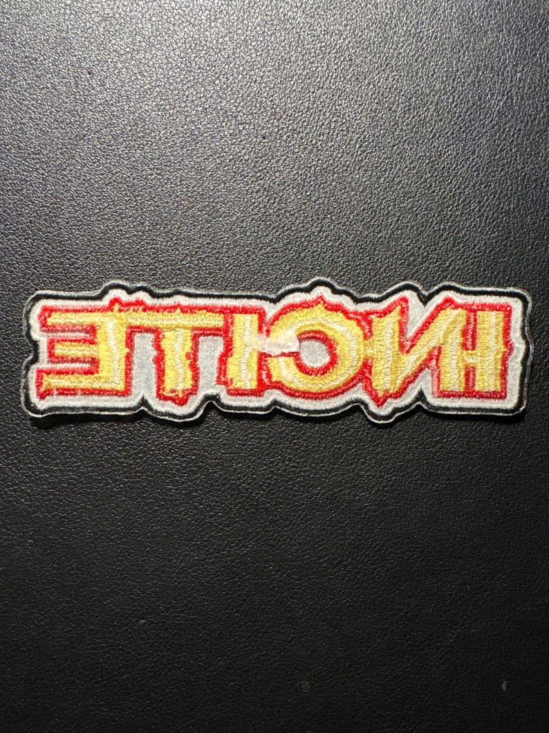 Op de afbeelding: Geborduurde patch met het woord "HUSTLE" in geel, omrand met rood en een witte rand. De patch heeft een rechthoekige vorm en is geplaatst tegen een zwarte achtergrond. De tekst is in een vet, gestileerd lettertype.