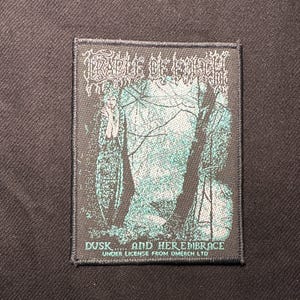 Könnte beinhalten: Ein schwarzer Aufnäher mit dem Bandnamen "Cradle of Filth" in weißen gotischen Buchstaben. Der Aufnäher hat eine türkis-schwarze Illustration mit dem Text "Dusk... and Her Embrace" und "Under license from OMERCH LTD."