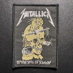 Könnte beinhalten: Schwarzer rechteckiger Aufnäher mit dem Bandnamen "METALLICA" in Silber, über einer goldenen Totenkopf-Grafik. Der Text "HARVESTER OF SORROW" steht darunter. Der Aufnäher hat einen schwarzen Rand.
