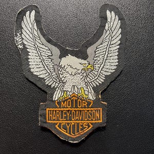 Può includere: Una toppa ricamata nera raffigurante un'aquila bianca con le ali spiegate sopra un emblema rettangolare arancione e nero. L'emblema mostra il testo MOTOR HARLEY-DAVIDSON CYCLES in lettere arancioni, con l'aquila dotata di un becco giallo.