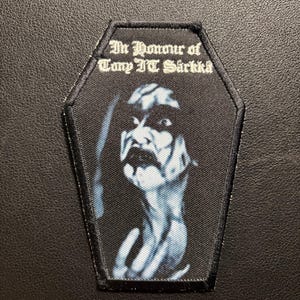 May include: A coffin-shaped embroidered patch with a black border. The centre features a high-contrast, blue-toned portrait of a person with long hair. Above the portrait, the text reads In Honour of Tony IT Särkkä in a gothic font.