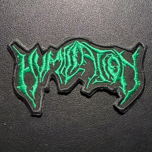 Op de afbeelding: Geborduurde patch met het woord "HUMIDITY" in een gestileerd, groen lettertype. De patch heeft een zwarte rand en is geplaatst tegen een donkere, getextureerde achtergrond. De belettering heeft een gothic- of metalband-esthetiek.