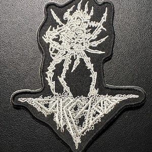 以下が含まれることがあります： 白黒の刺繍パッチで、骸骨のような生き物のデザインが特徴です。 パッチはユニークな形で、骸骨の姿と「SARCOMA」という言葉が白で詳細に描かれています。